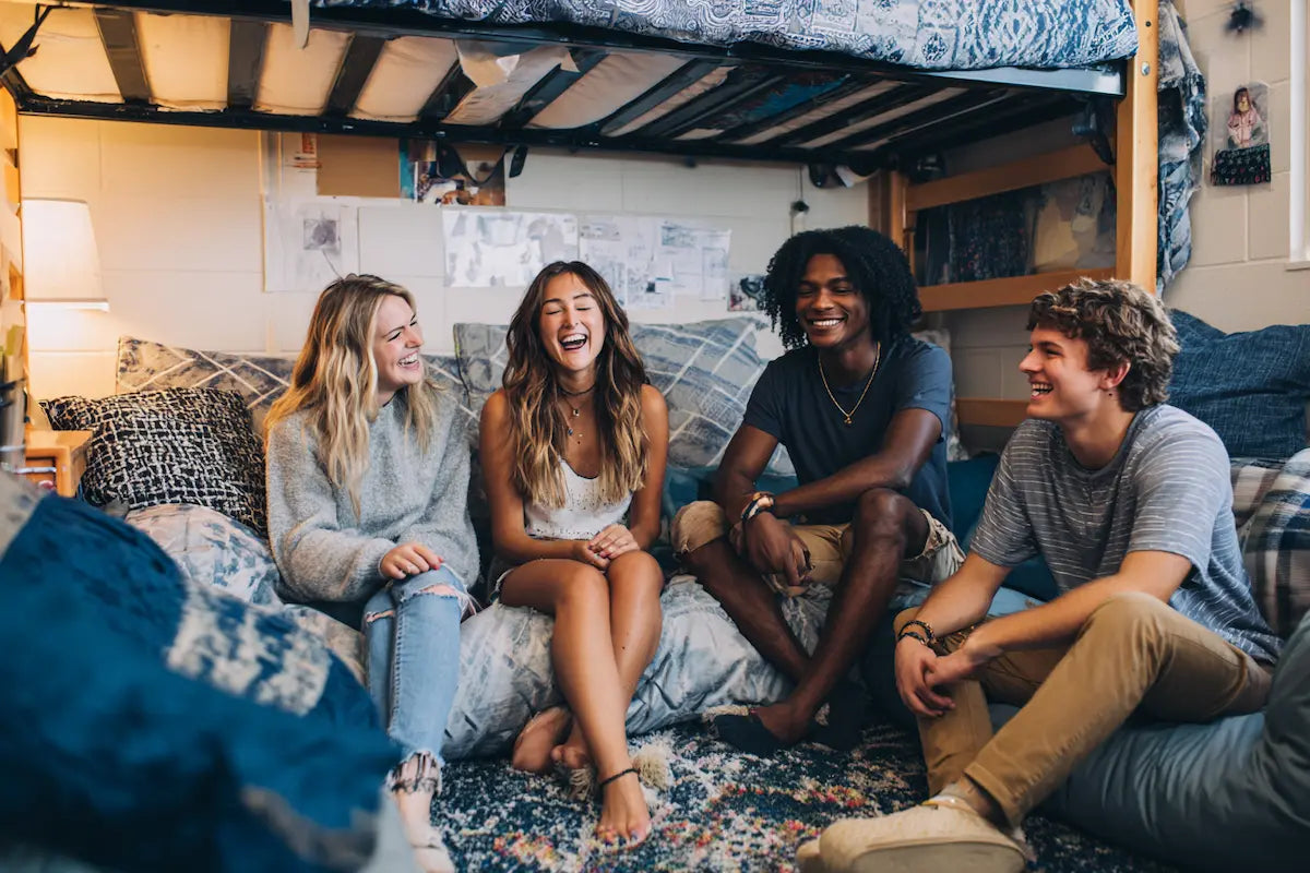 Four college students laughing and sitting on a futon in a dorm room with a blue color palette